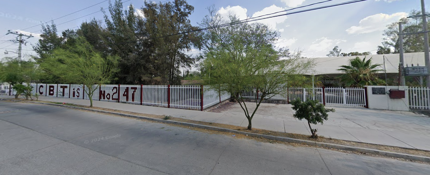 Fotografía en la que se muestra una explanada. Al terminar la explanada hay una reja blanca que da acceso a las instalaciones. El plantel está rodeado por rejas blancas y encima de estas rejas, están pintadas letras rojas que forman la palabra Cebetis número 247.